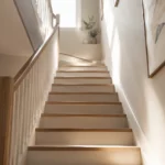 Habiller une cage d'escalier : la méthode pour transformer un couloir en circulation qui a du style Cage d'escalier intérieure contemporaine avec murs clairs, rampe en bois et rai de lumière naturelle illuminant les premières marches.