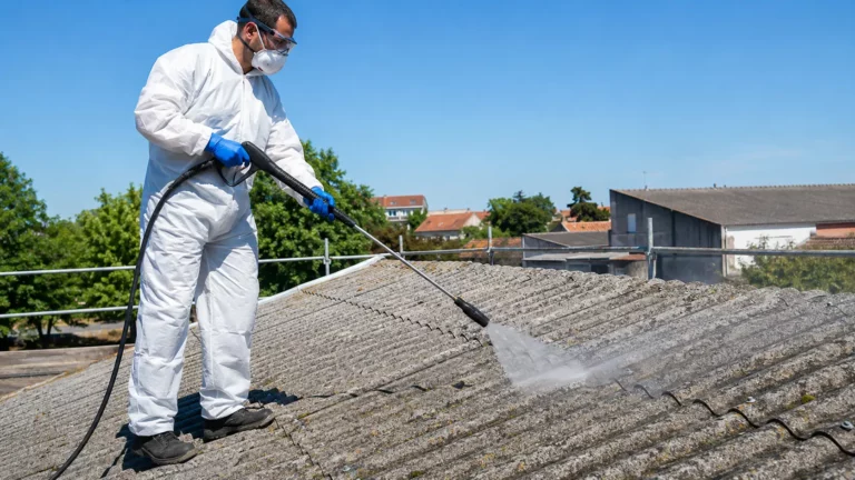 Professionnel du bâtiment équipé d'une combinaison jetable, d'un masque FFP3, de gants nitrile et de lunettes de protection nettoyant une toiture en fibrociment avec un nettoyeur basse pression à lance à jet plat, en maintenant une distance de sécurité sur un chantier résidentiel.