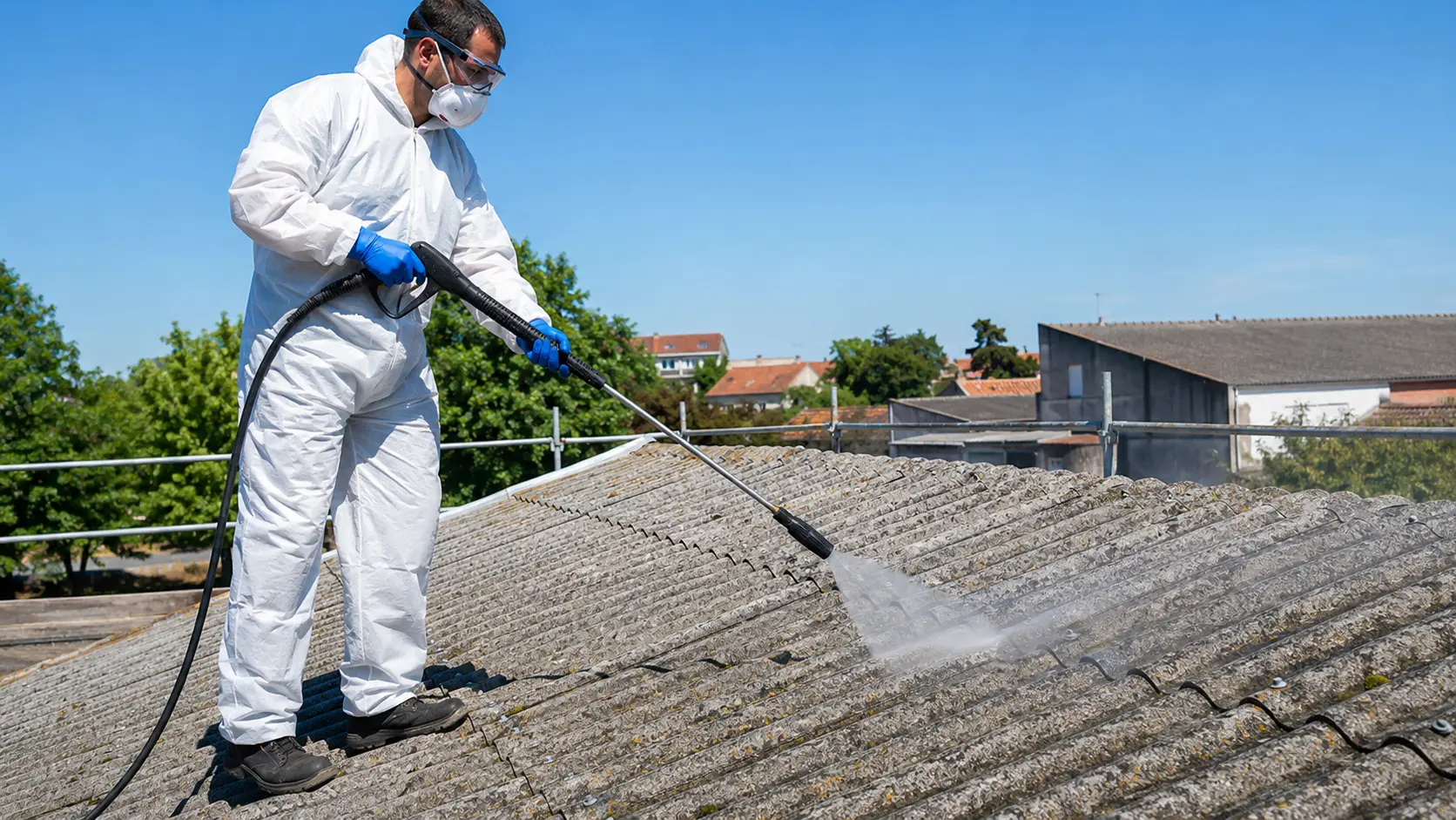 Nettoyage toiture fibrociment : la méthode qui protège votre toit (et vos poumons) Professionnel du bâtiment équipé d'une combinaison jetable, d'un masque FFP3, de gants nitrile et de lunettes de protection nettoyant une toiture en fibrociment avec un nettoyeur basse pression à lance à jet plat, en maintenant une distance de sécurité sur un chantier résidentiel.