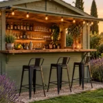 Abri de jardin en bois vert sauge aménagé en bar extérieur avec comptoir, tabourets hauts et guirlandes lumineuses, entouré de lavande.